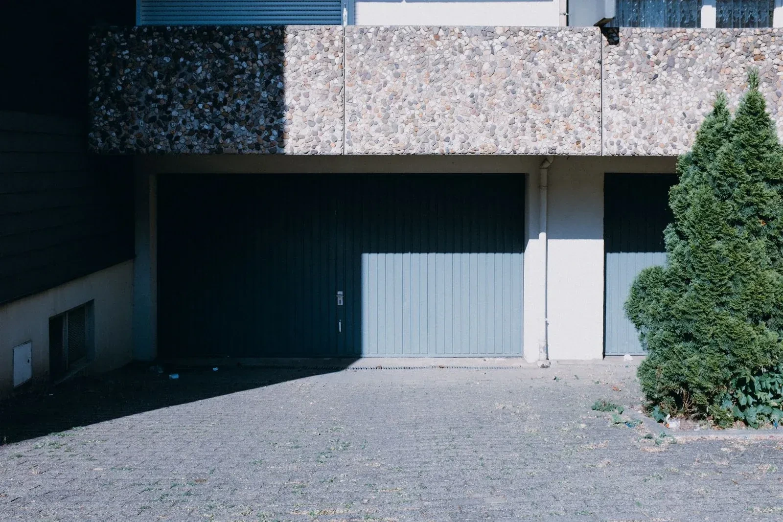 Two gray-blue vertical-plank garage doors under a pebble-textured concrete overhang, half in shadow, beside a green shrub and a paved lot.